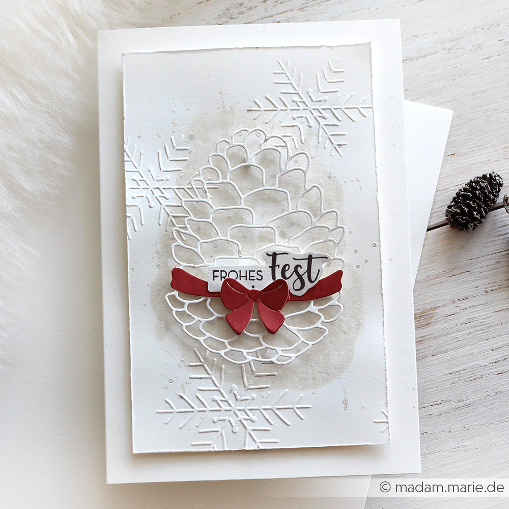 Handgemachte Weihnachtskarte von Alexandra Renke mit Motivtannenzapfen, roter Schleife und "Frohes Fest" auf weißem Designpapier mit geprägten Schneeflocken.