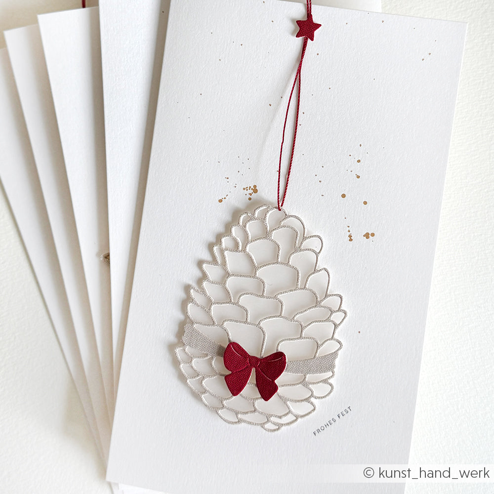 Weiße Weihnachtskarte von Alexandra Renke aus Designpapier mit einem Tannenzapfen aus der Motivstanze Geschenkzapfen, der mit einer roten Schleife, einer roten Schnur und einem Stern verziert ist und auf anderen Karten liegt.
