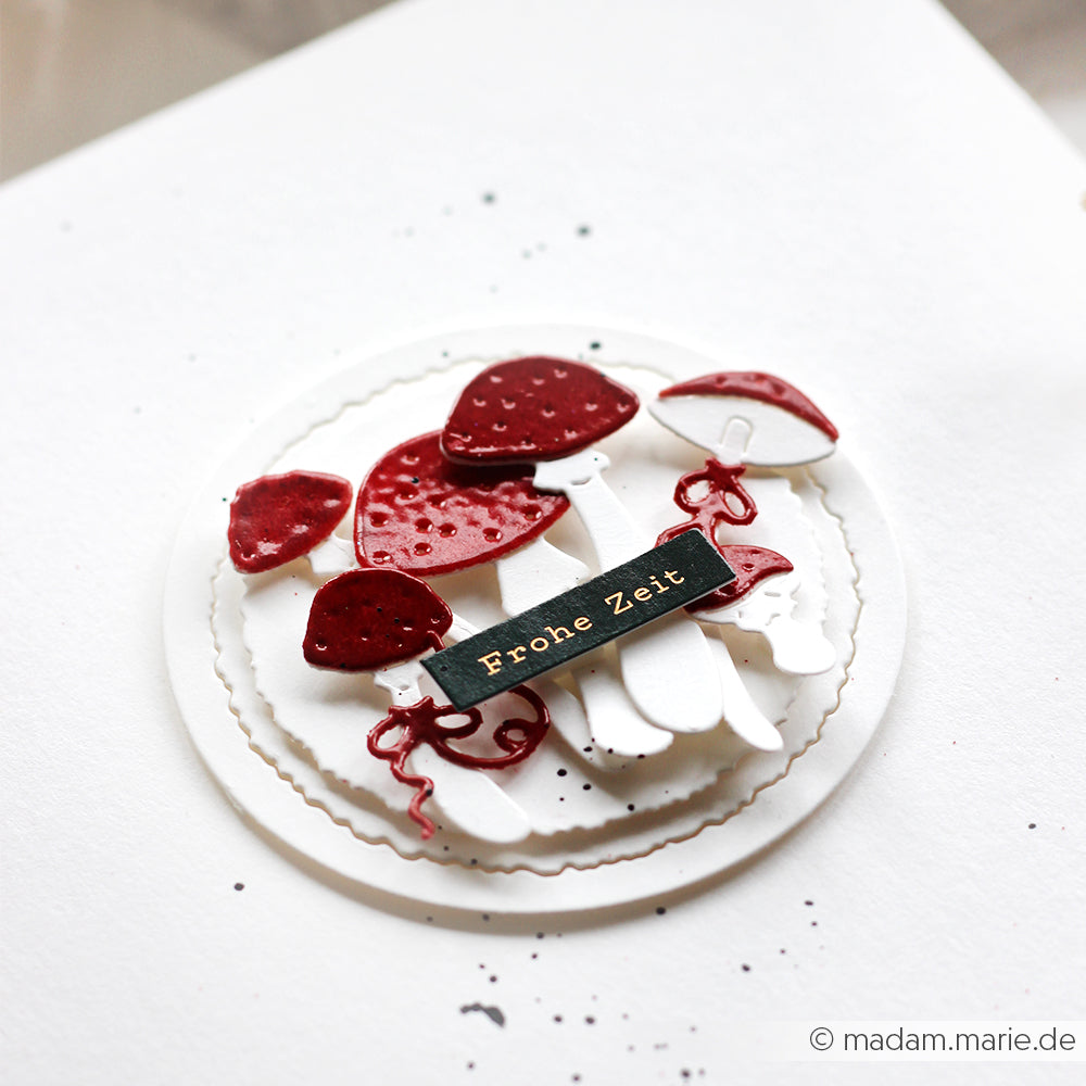 Eine runde Karte mit roten und weißen Pilzen, die mit dem Motivstanze Glückspilze-Trio von Alexandra Renke hergestellt wurde, zeigt ein "Frohe Zeit"-Label auf weißem Hintergrund.