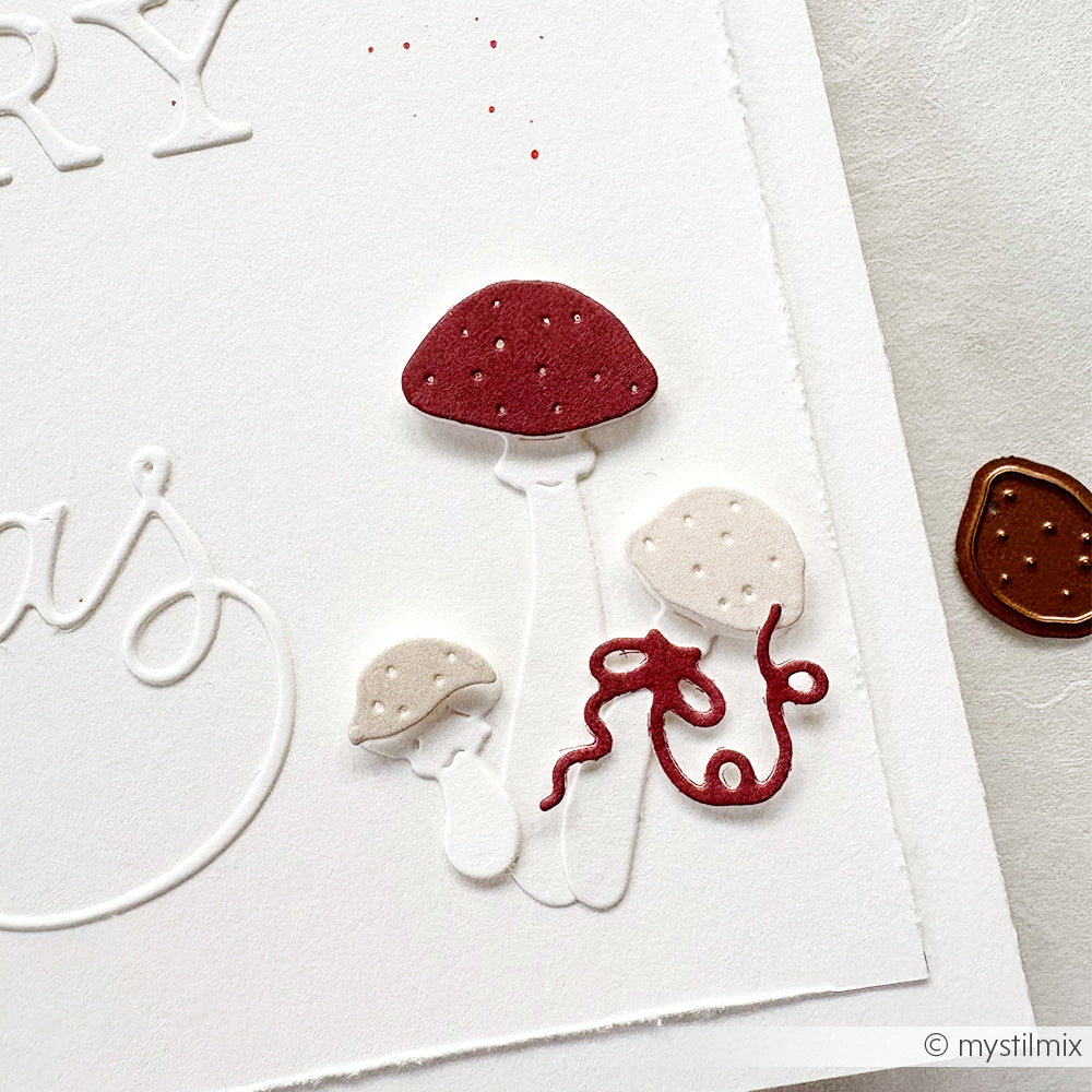 Weiße Karte mit geprägten Buchstaben und drei strukturierten Pilzen - zwei weiße und ein roter - sowie einem roten Wirbel, der mit der Motivstanze Glückspilze von Alexandra Renke hergestellt wurde und für eine charmante Note sorgt.