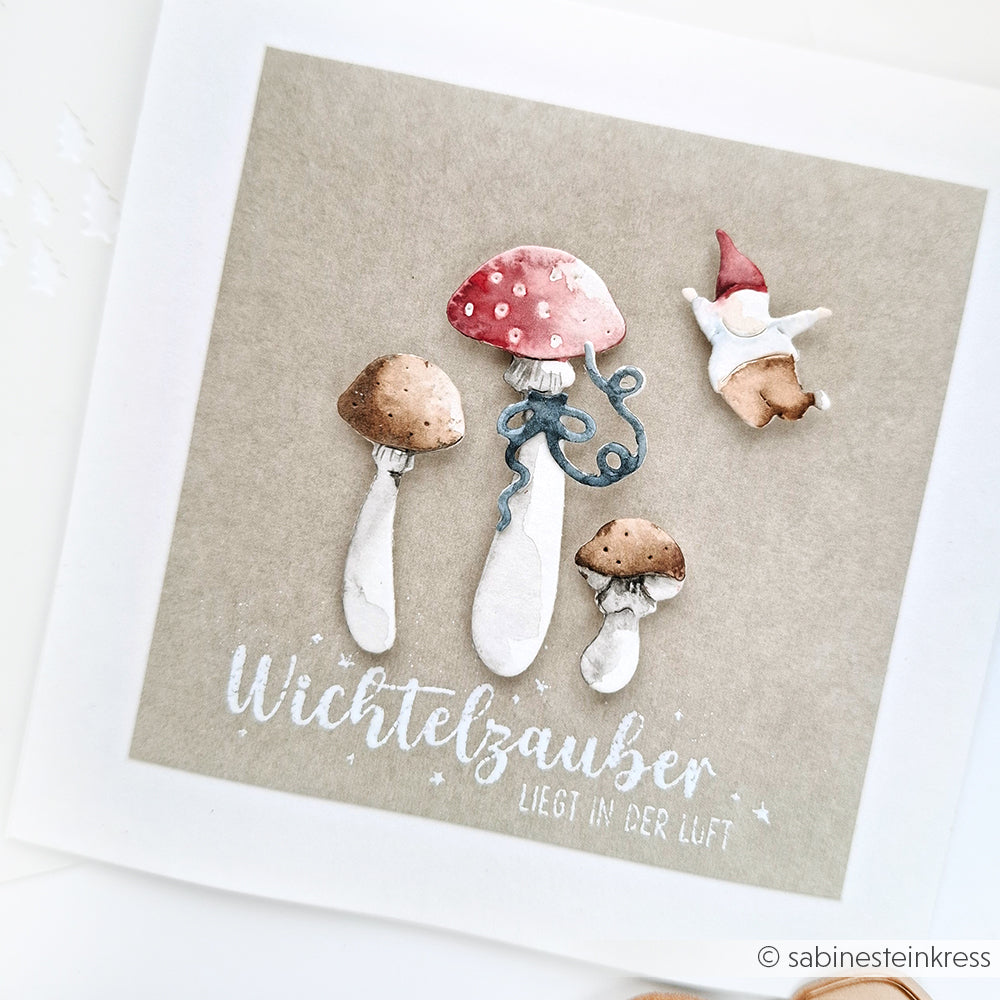 Aquarellillustration von drei Pilzen und einem kleinen Zwerg mit dem Text "Wichtelzauber liegt in der Luft". Perfekt für Scrapbooking oder das Glückspilze-Trio-Stanzsystem von Alexandra Renke.