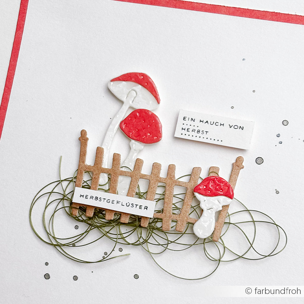 Handgefertigte Karte mit roten Pilzen, die mit der Glückspilze-Trio-Stanze von Alexandra Renke, einem braunen Zaun, grünen Fäden und einem deutschen Herbsttext auf weißem Designpapier hergestellt wurde.