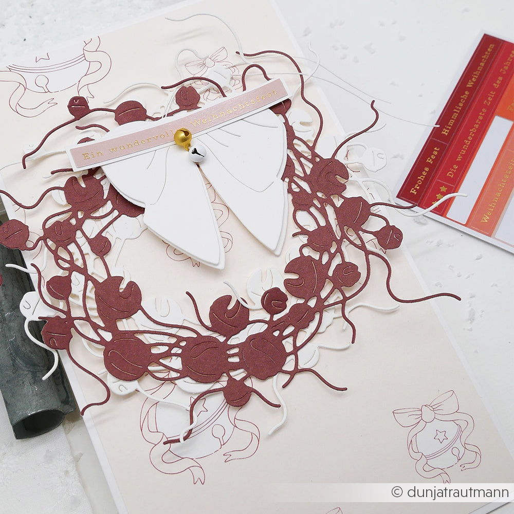 Die handgefertigte Karte von Alexandra Renke zeigt einen weinroten und weißen Motivstanze Glöckchenkranz, eine weiße Schleife und eine goldene Spange auf hellem Hintergrund, die mit eleganten Designpapieren verziert ist.