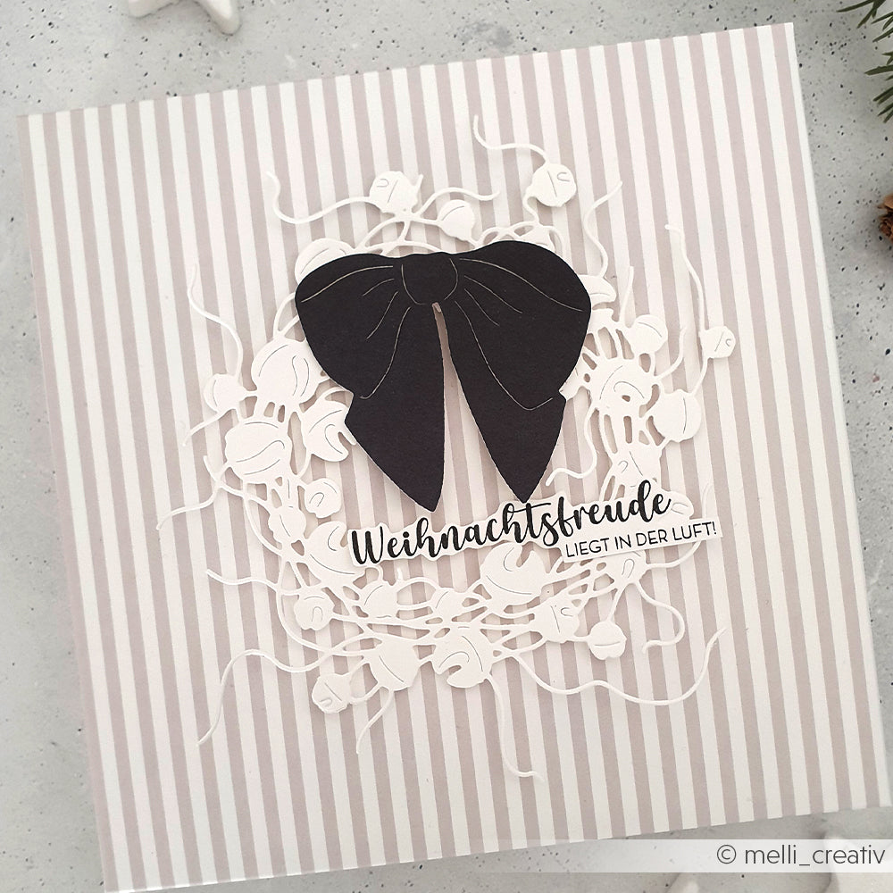 Quadratische Karte von Alexandra Renke mit einem weißen Kranz aus der Motivstanze Glöckchenkranz, akzentuiert mit einer schwarzen Schleife und deutschem Text, auf gestreiftem Designpapier.
