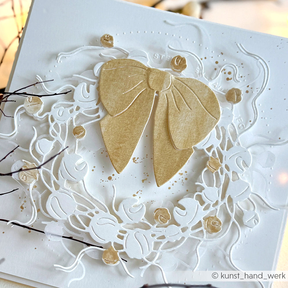 Eine weiße Prägekarte von Alexandra Renke mit einer goldenen Schleife und einem Blumenkranz, die mit der Motivstanze Glöckchenkranz und elegantem Designpapier hergestellt wurden.