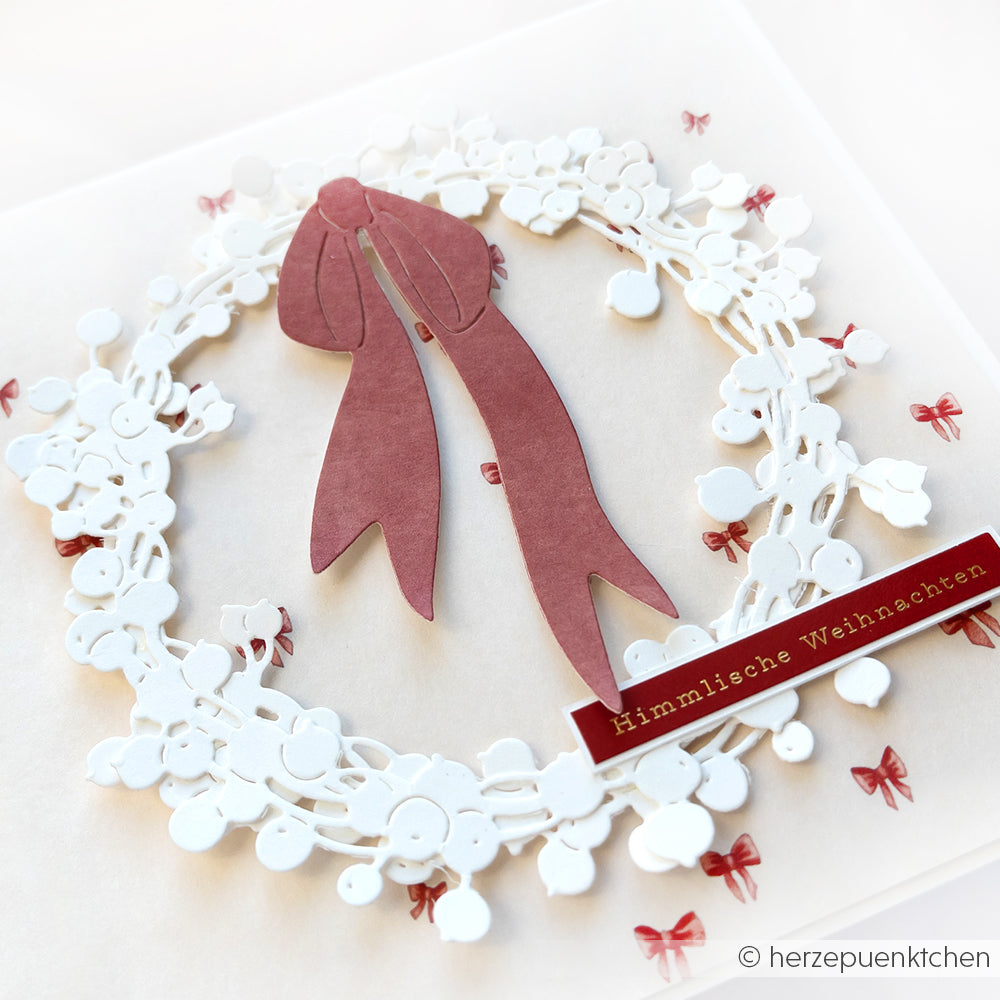 Eine festliche Karte mit einem weißen Blumenkranz aus der Alexandra Renke Motivstanze Großer Beerenkranz, einer roten Schleife und dem Text "Himmlische Weihnachten" auf hochwertigen Designpapieren.