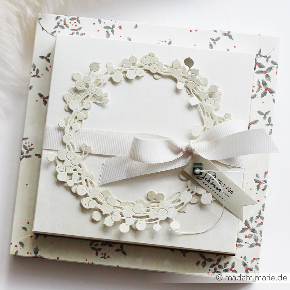 Diese handgefertigte weiße Karte von Alexandra Renke zeigt einen Papierkranz aus der Motivstanze Großer Beerenkranz, Schleifenband und ein kleines Schildchen, schön präsentiert auf einem Umschlag, der mit exquisiten floralen Designpapieren verziert ist.
