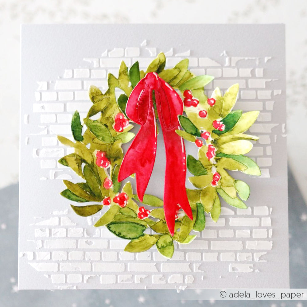 Handgefertigte Karte mit einem aquarellierten Kranz mit roter Schleife und Beeren auf einem weißen Hintergrund mit Ziegelsteinmuster, hergestellt mit Alexandra Renke Designpapieren und Motivstanze Blätterkranz DieCut Systeme.