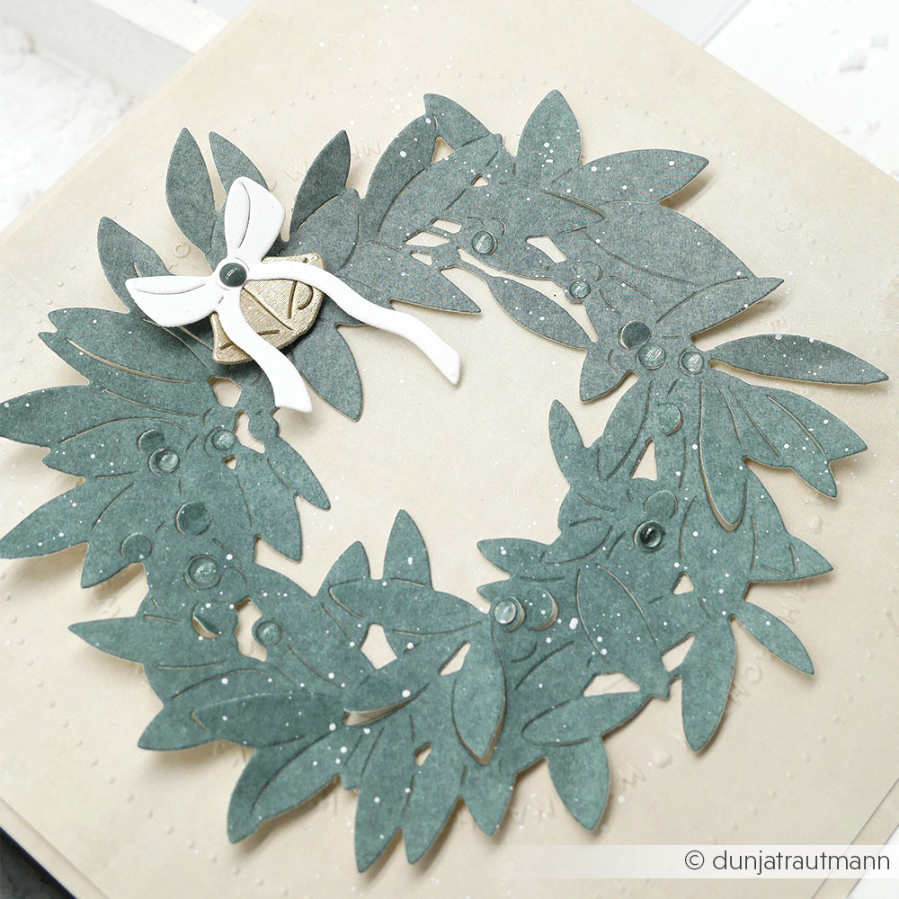 Ein Papierkranz mit grünen Blättern aus der Motivstanze Blätterkranz von Alexandra Renke, verziert mit einer kleinen weißen Schleife und zarten weißen Sprenkeln, vor einem beigen Hintergrund.