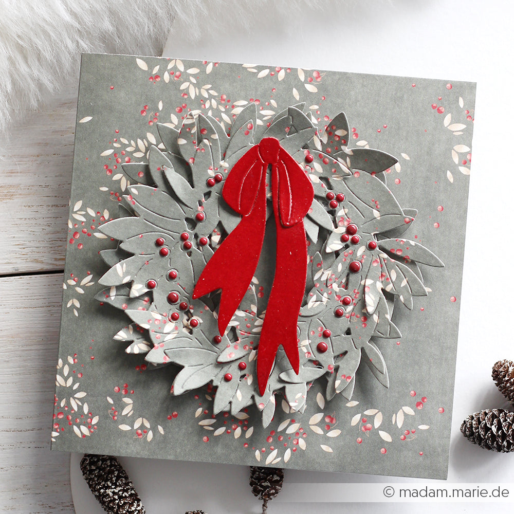 Eine graue Weihnachtskarte mit einem Blätterkranz aus der Alexandra Renke Motivstanze Blätterkranz, verziert mit roten Beeren und einer roten Schleife aus Band und umgeben von Tannenzapfen.