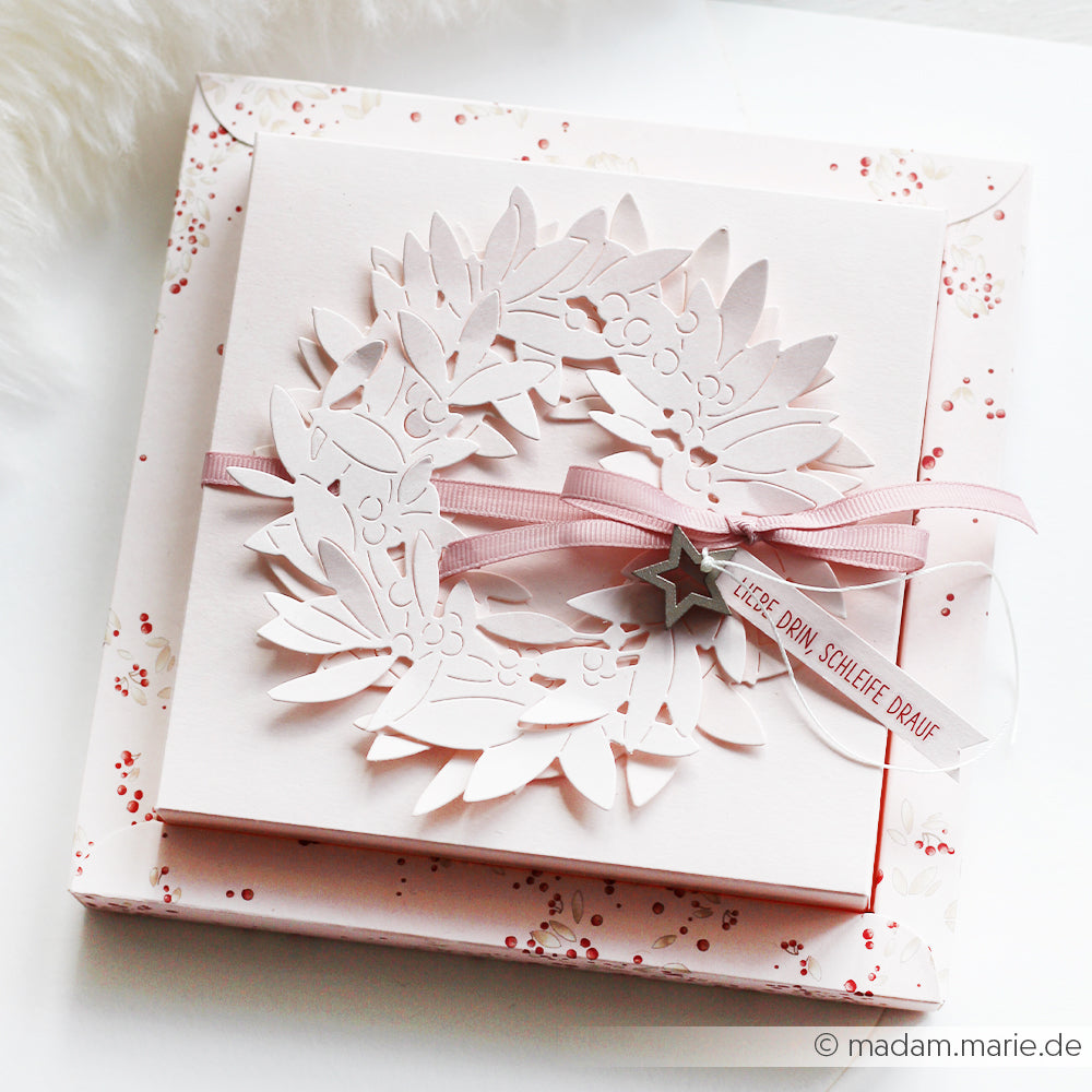 Viereckige pastellfarbene Geschenkbox von Alexandra Renke mit einem Papierkranz aus Motivstanze Blätterkranz, rosa Schleife und einem kleinen Anhänger auf einem blumengemusterten Deckel.