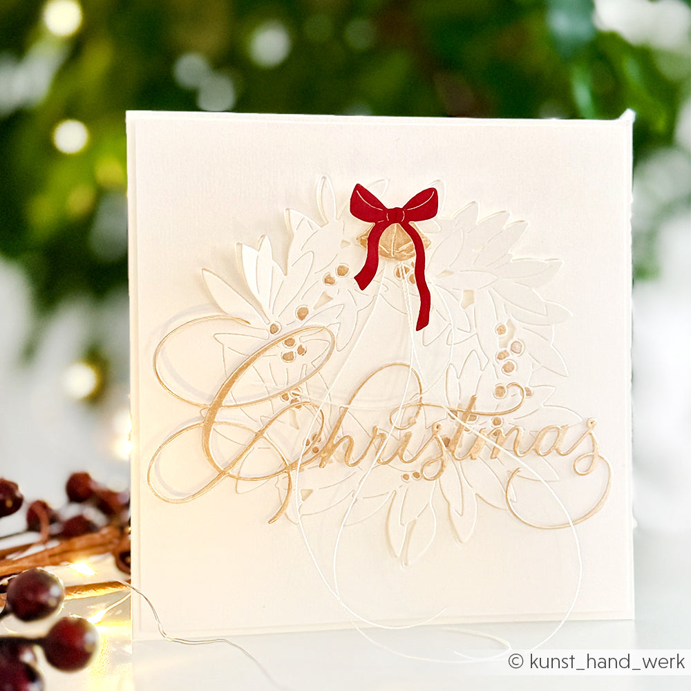 Weiße Weihnachtskarte von Alexandra Renke mit goldener Schrift, Akzenten aus Designpapier, weißen Blättern wie eine Motivstanze Blätterkranz und einer roten Schleife; verschwommene festliche Lichter im Hintergrund.