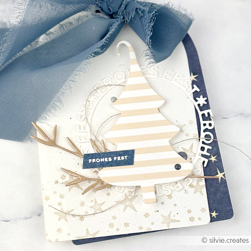 Gestreifte Papier-Weihnachtsbaumkarte mit der Motivstanze Fröhliche Tanne von Alexandra Renke, mit deutschem Text, blauem Band, goldenen Sternen und festlichen Verzierungen - ideal für kreative DieCut Systeme Projekte.