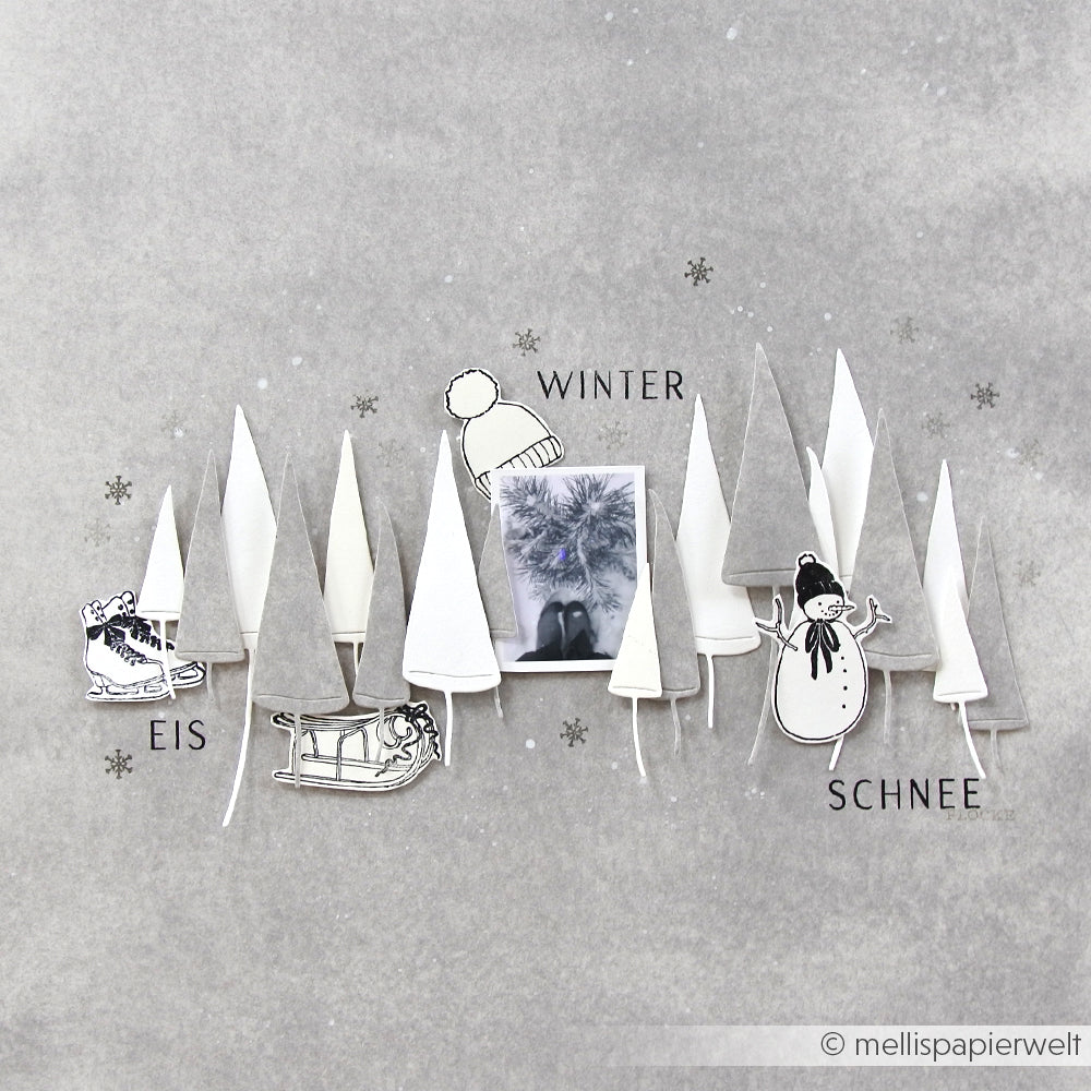 Mit Alexandra Renke Designpapiere und DieCut Systeme gestaltete Papierbäume sowie ein Schlitten, ein Schneemann und eine Wintermütze umrahmen ein Foto auf einem strukturierten grauen Hintergrund. Motivstanze Schiefe Tannen verleiht dieser gemütlichen Winterszene Charme.