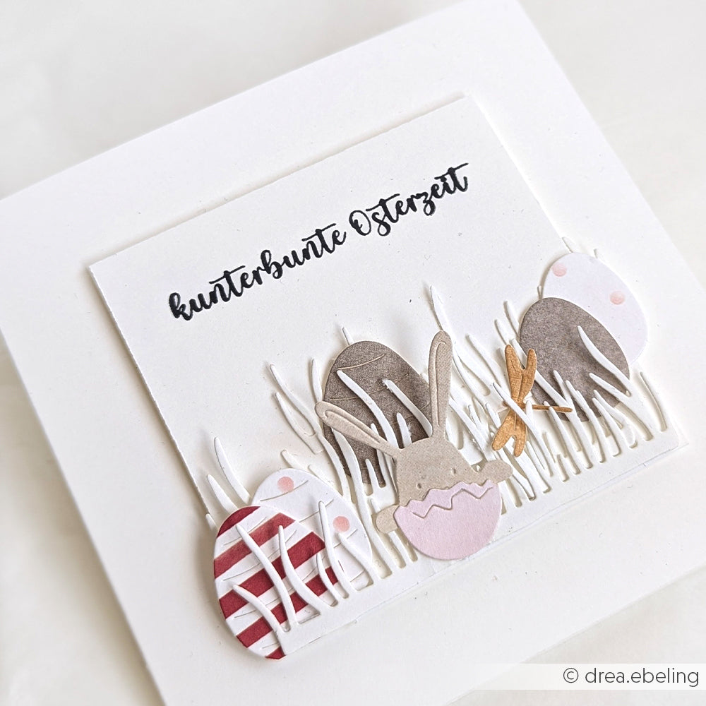Die handgefertigte Karte von Alexandra Renke zeigt pastellfarbene Eier, einen Hasen und Gras, hergestellt mit Designpapieren und dem Motivstanze Grasbüschel-Set, veredelt mit dem Text "kunterbunte Osterzeit" über dem Motiv.