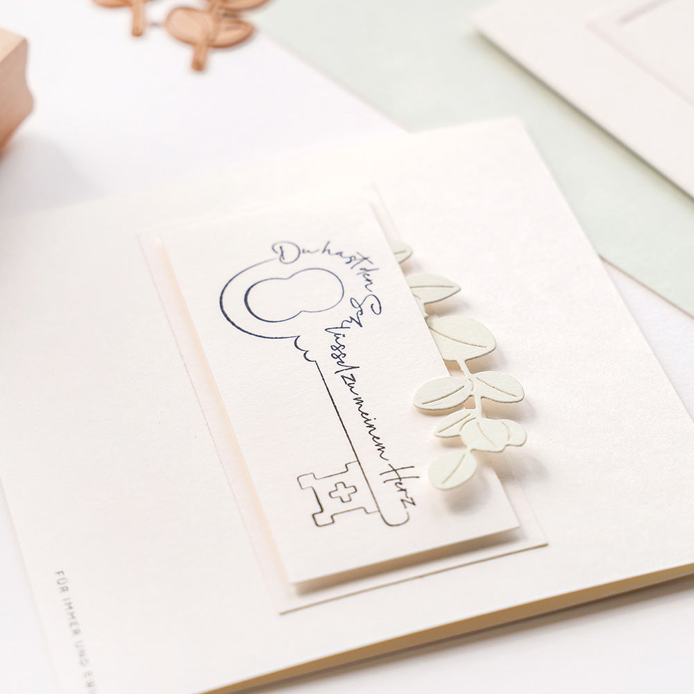 Eine weiße Karte von Alexandra Renke mit einer Schlüsselillustration, handgeschriebener Schrift und Blattverzierungen aus Motivstanze Eukalyptuszweige auf Designpapieren vor einem hellen Hintergrund.
