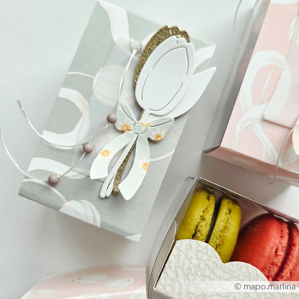 Eine dekorative Geschenkschachtel von Alexandra Renke mit einem weißen Blumenband neben einer offenen Schachtel mit gelben und roten Macarons, hergestellt aus hochwertigen Designpapieren und veredelt mit DieCut Systemen für ein elegantes Finish.