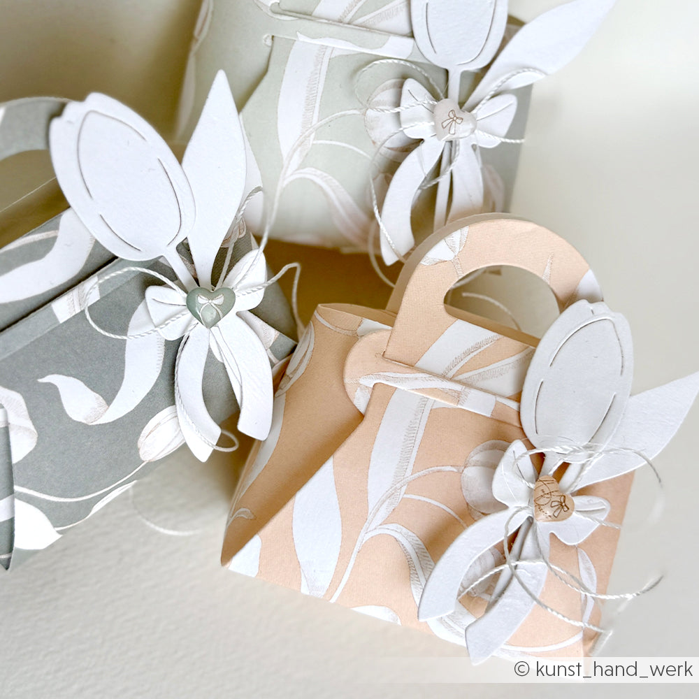 Drei handtaschenförmige Geschenkboxen aus Alexandra Renke Designpapieren, verziert mit weißen Blumenausschnitten mit Stanzsystemen und der Motivstanze Tulpensträußchen für einen eleganten Touch.