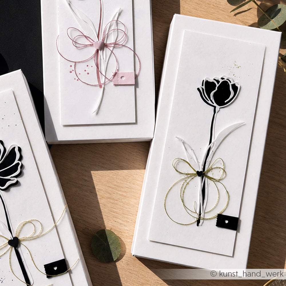 Drei rechteckige, handgefertigte Karten von Alexandra Renke mit Blumen- und Schleifenmotiven, hergestellt mit Designpapieren und DieCut Systemen in Schwarz, Weiß, Rosa und Gold, wunderschön arrangiert auf einer Holzoberfläche.