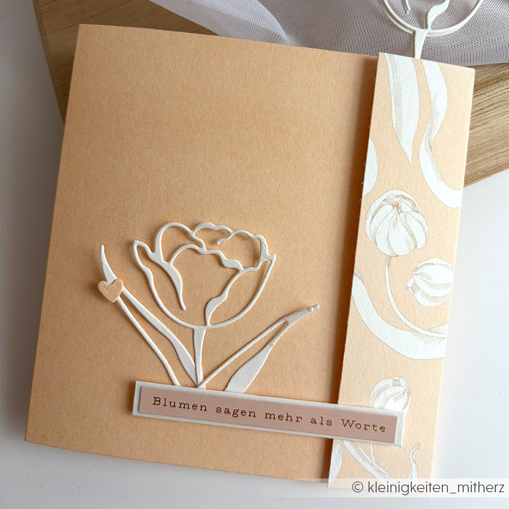 Pfirsichfarbene Karte von Alexandra Renke mit einer weißen Tulpe und dem deutschen Text "Blumen sagen mehr als Worte". Hergestellt mit Motivstanze Lineart Kelchblume-perfekt für einzigartiges Designpapier oder die Verwendung mit DieCut Systemen.
