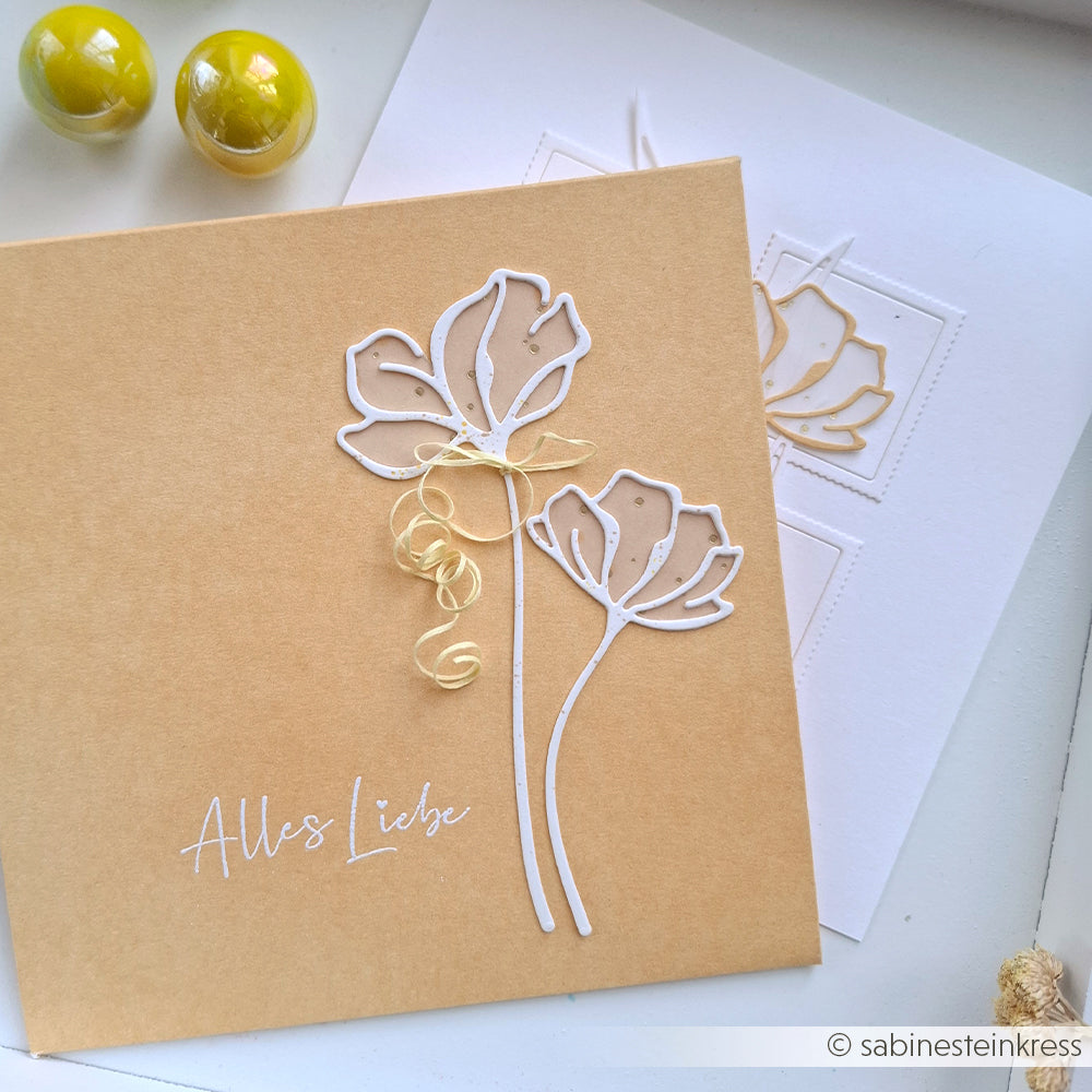 Quadratische Karte mit zwei weißen Blumenumrissen, die mit der Alexandra Renke Motivstanze Lineart Fächerblumen und dem Text "Alles Liebe" hergestellt wurden, neben einer weißen Karte und gelben Kugeln.