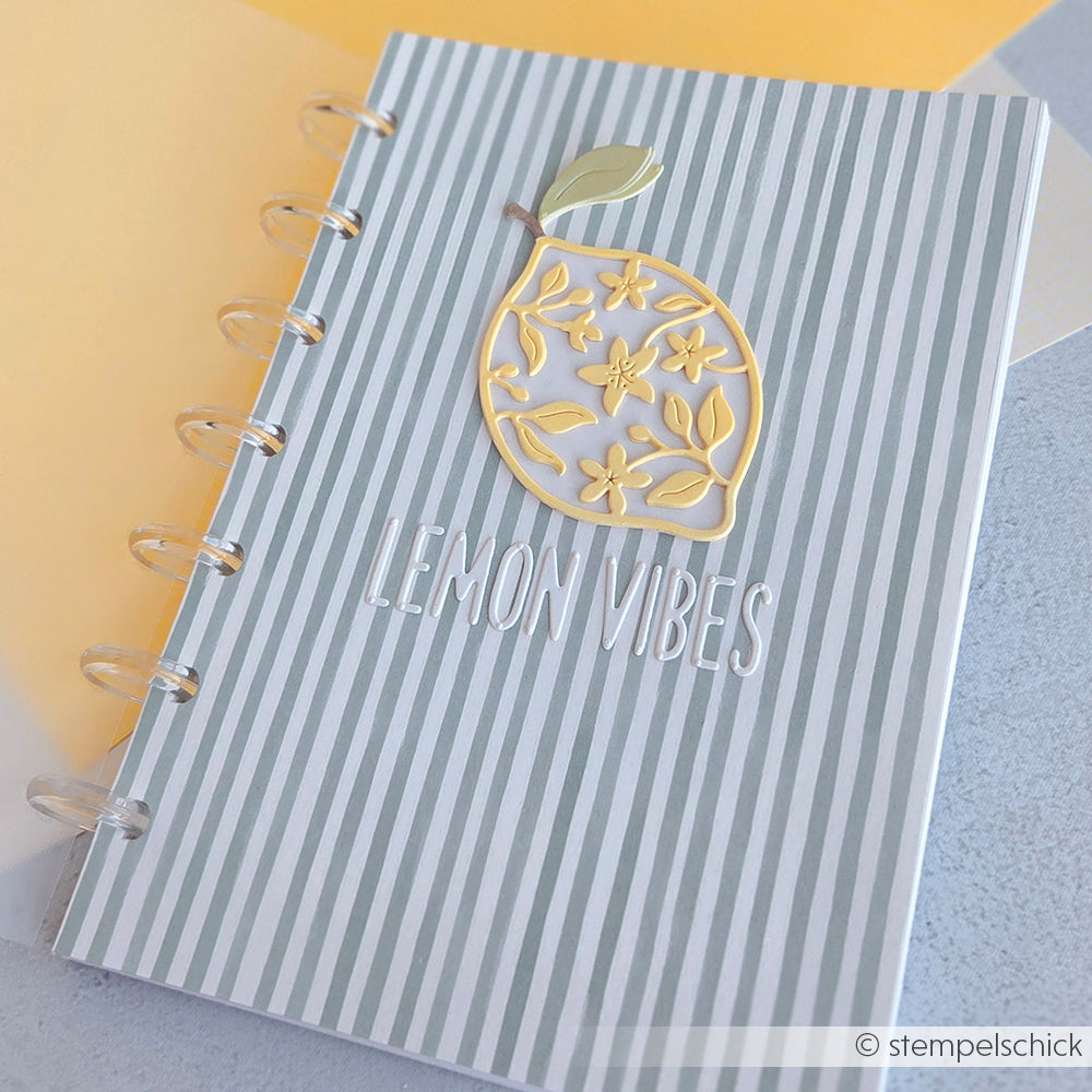 Notizbuch mit grauen und weißen Streifen, einem gelben Zitronenmotiv, inspiriert von der Motivstanze „Blumige Zitrone“, und dem Schriftzug „LEMON VIBES“ auf dem Einband.
