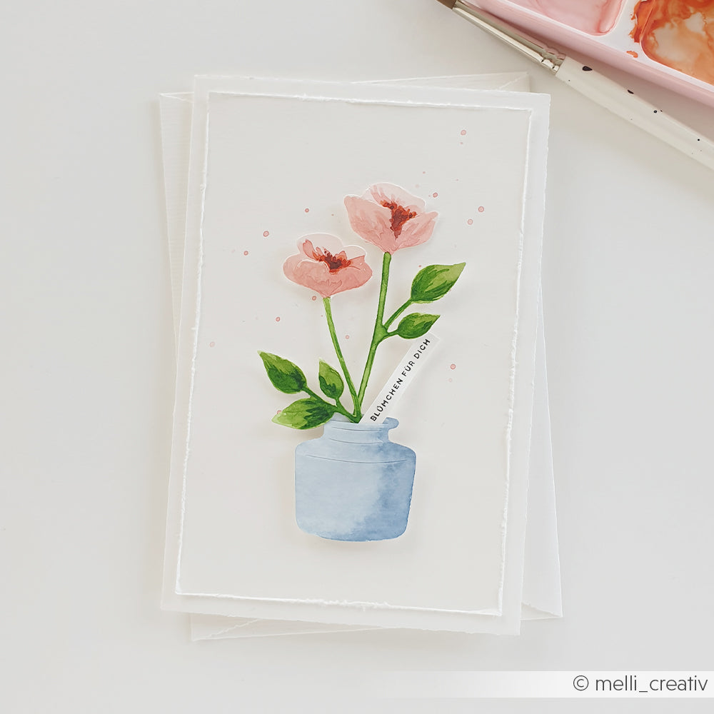 Aquarellillustration von zwei rosa Blumen in einer blauen Vase auf einer weißen Grußkarte von Alexandra Renke, mit Malutensilien und Stanzsystemen, die dieses von der Motivstanze „Blütenpoesie“ inspirierte Design unterstreichen.