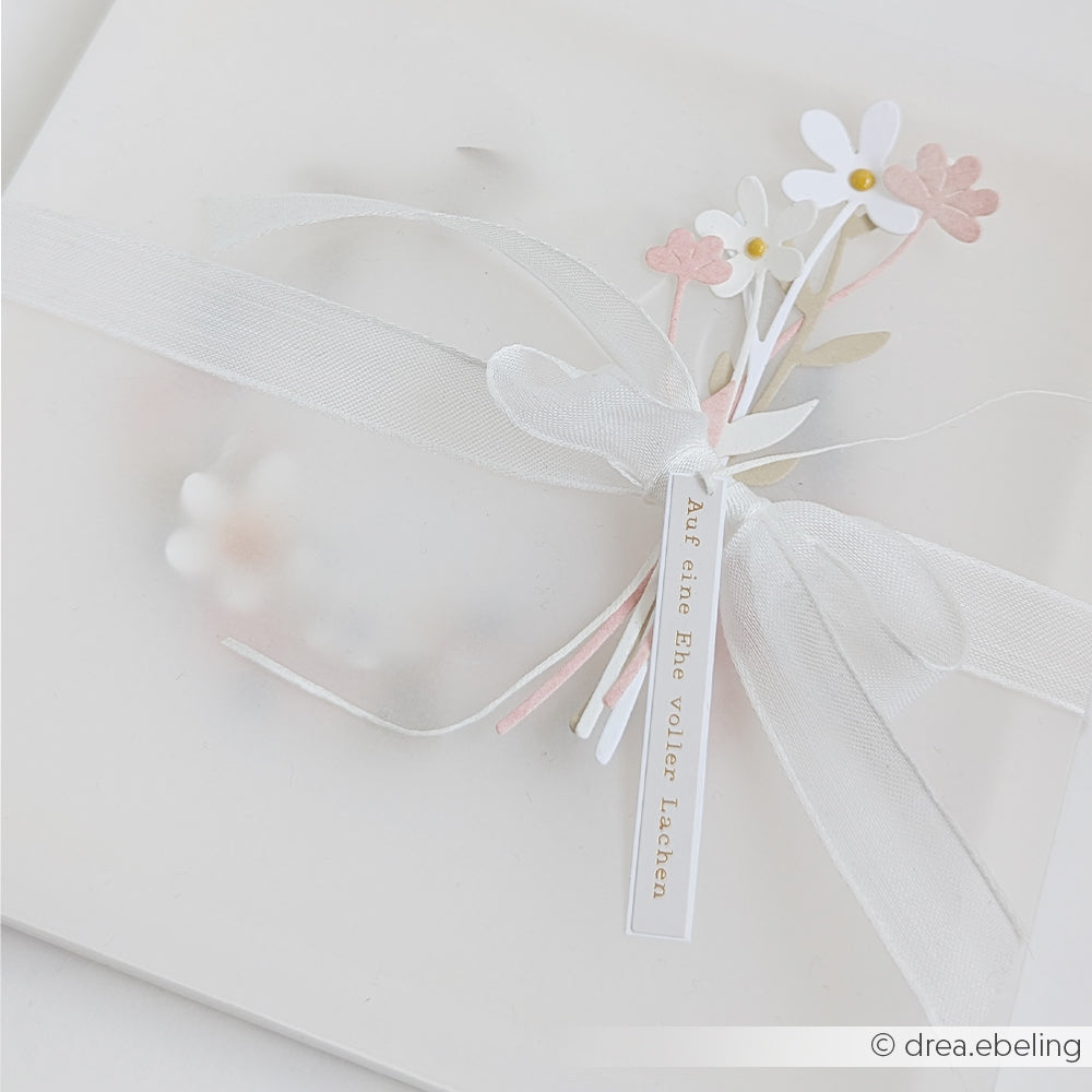 Quadratische weiße Karte von Alexandra Renke, verziert mit einem transparenten Band, Papierblumen, die mit der Motivstanze „Zarte Blüten“ und hochwertigen Designpapieren gestaltet wurden, sowie einem kleinen Textanhänger auf Deutsch.