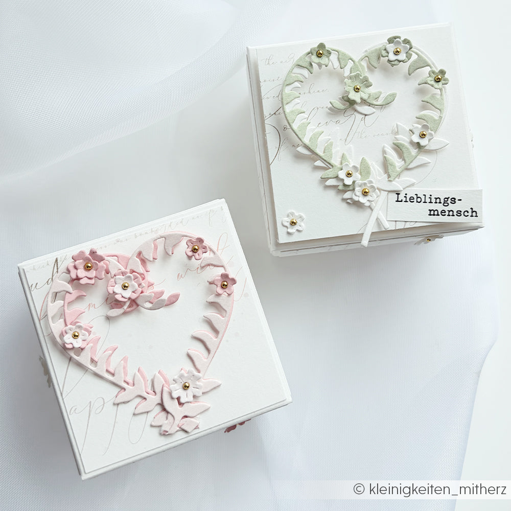 Zwei weiße Geschenkboxen mit dekorativen Herz- und Blumenmotiven, eine in Rosa und eine in Grün, die mit Stanzsystemen und Stanzformen für Designpapier gefertigt wurden und elegant auf einem weißen Untergrund präsentiert werden.