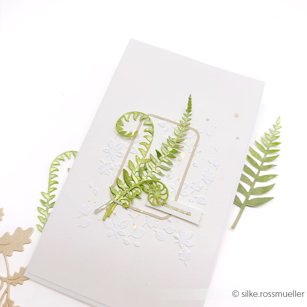 Eine weiße Grußkarte mit strukturierten Designpapieren und ausgeschnittenen grünen Farnblättern, die mit der Motivstanze „Farn Paar“ von DieCut Systeme gestaltet und kunstvoll auf der Vorderseite angeordnet wurden.