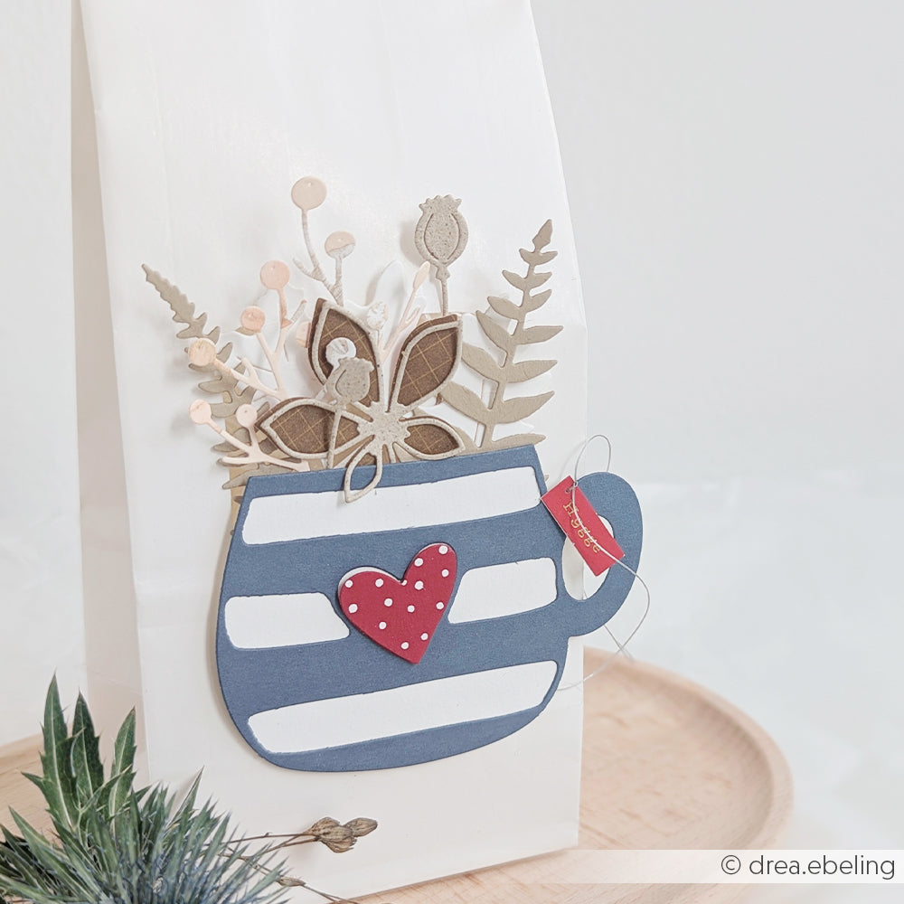 Geschenktüte mit einer blau gestreiften Tasse, einem roten Herz und einer Dekoration aus Trockenblumen auf der Vorderseite, wunderschön akzentuiert mit den „Farnzweige“-Stanzen von Motivstanze für Papier, die ihr eine elegante Note verleihen.