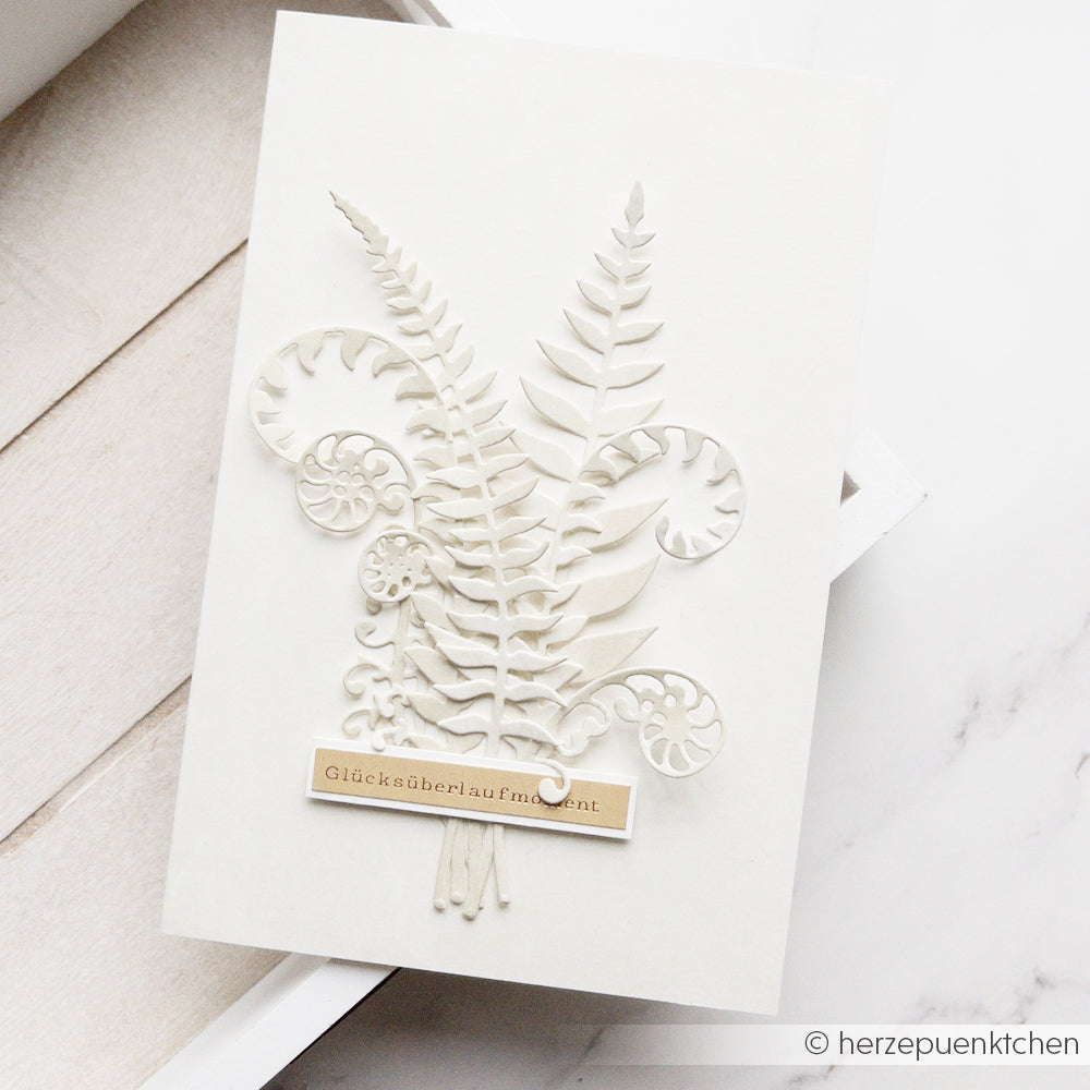 Weiße Karte mit geprägten Farnen, Blättern und einem kleinen Etikett mit der Aufschrift „Glücksüberlaufsmoment“, gefertigt mit der Motivstanze „Farn Spiralen“ und hochwertigen Designpapieren für ein edles Finish.