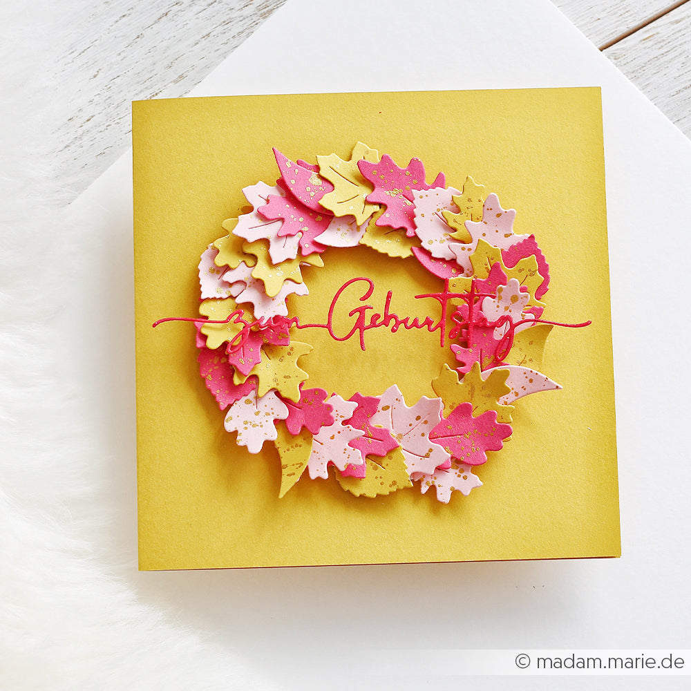 Gelbe Karte mit einem bunten Blattkranz aus Designpapier, gestaltet mit der Motivstanze „Herbstlaub“, in deren Mitte in roter Schreibschrift „Geburtstag“ steht.