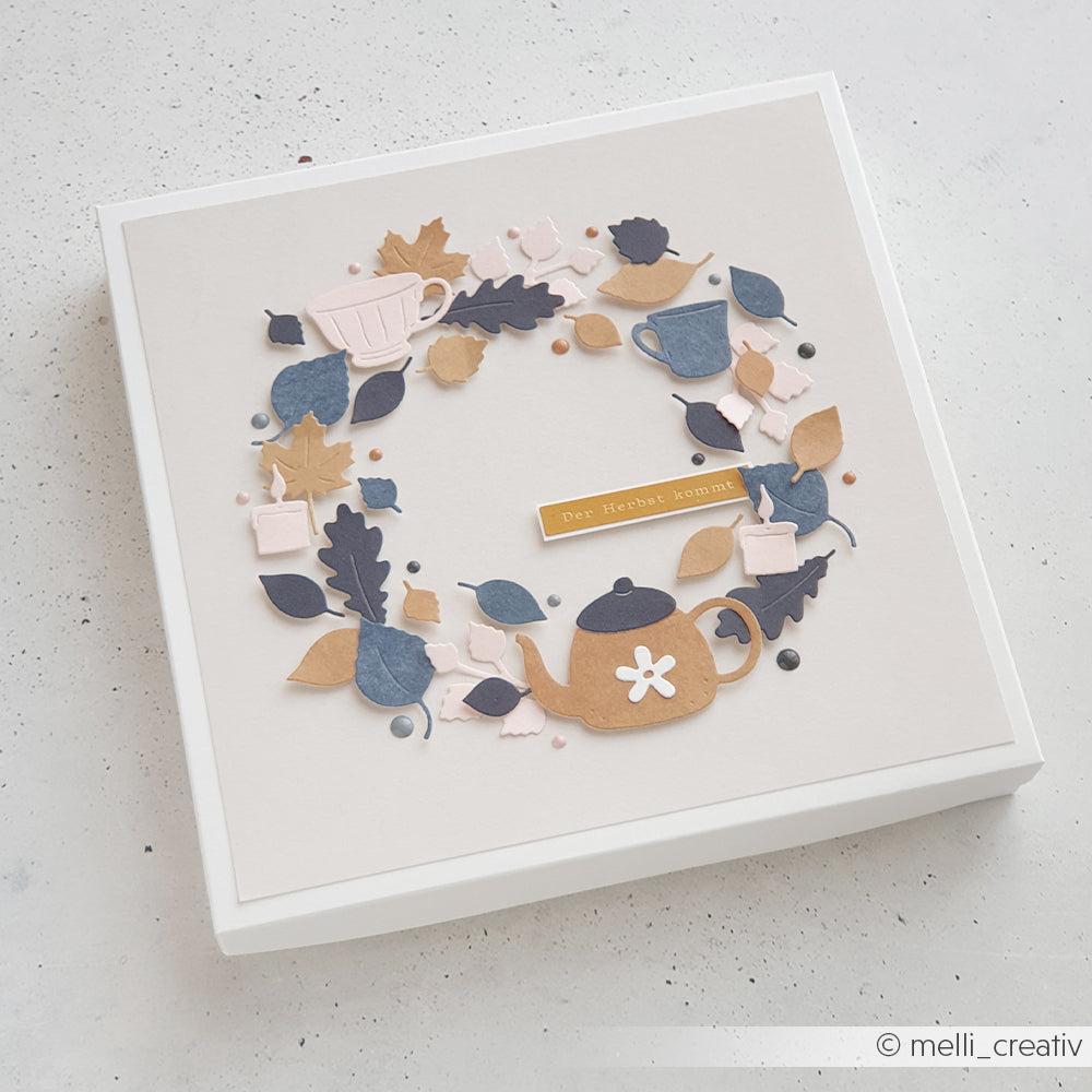 Ein Papierkranz aus Teekanne, Tassen und Herbstblättern auf einer beigen, quadratischen Kartonschachtel – er bringt die Designpapiere und die filigranen Details, die mit der Motivstanze „Herbstlaub“ von DieCut Systeme gestaltet wurden, perfekt zur Geltung.
