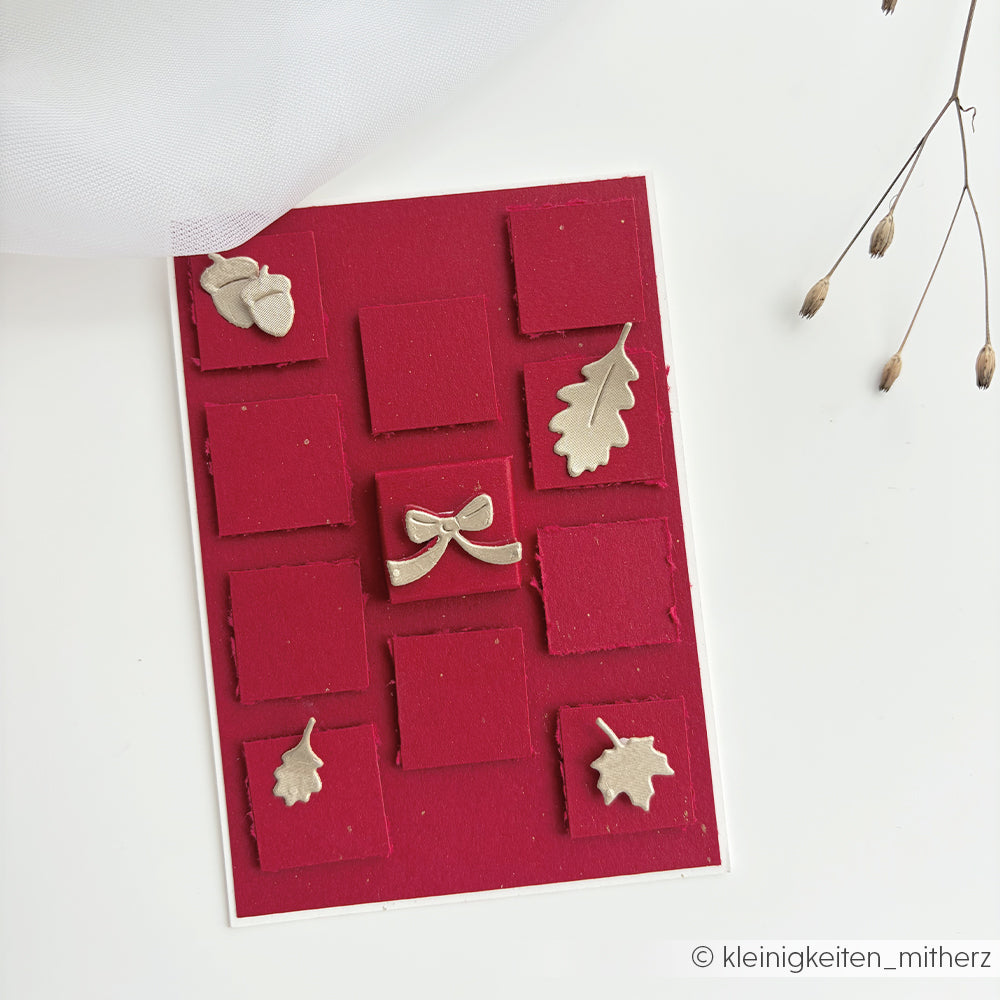 Rote Karte mit erhabenen Quadraten und Verzierungen aus Blattgold, Schleifen und Eicheln, gestaltet mit „Stanzen für Designpapier“ für zusätzliche Struktur und Details, auf weißem Hintergrund.