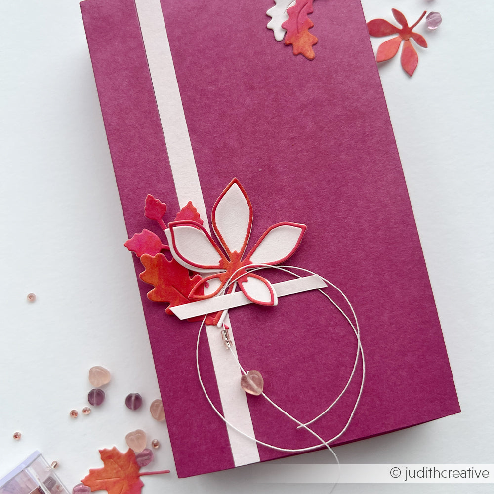 Lila Geschenkbox mit weißer Schleife, gefertigt aus eleganten Designpapieren, verziert mit Blumen- und Blattmotiven, die mit der Motivstanze „Blätterwirbel“ gestaltet wurden, und abgerundet mit Perlen auf hellem Hintergrund.