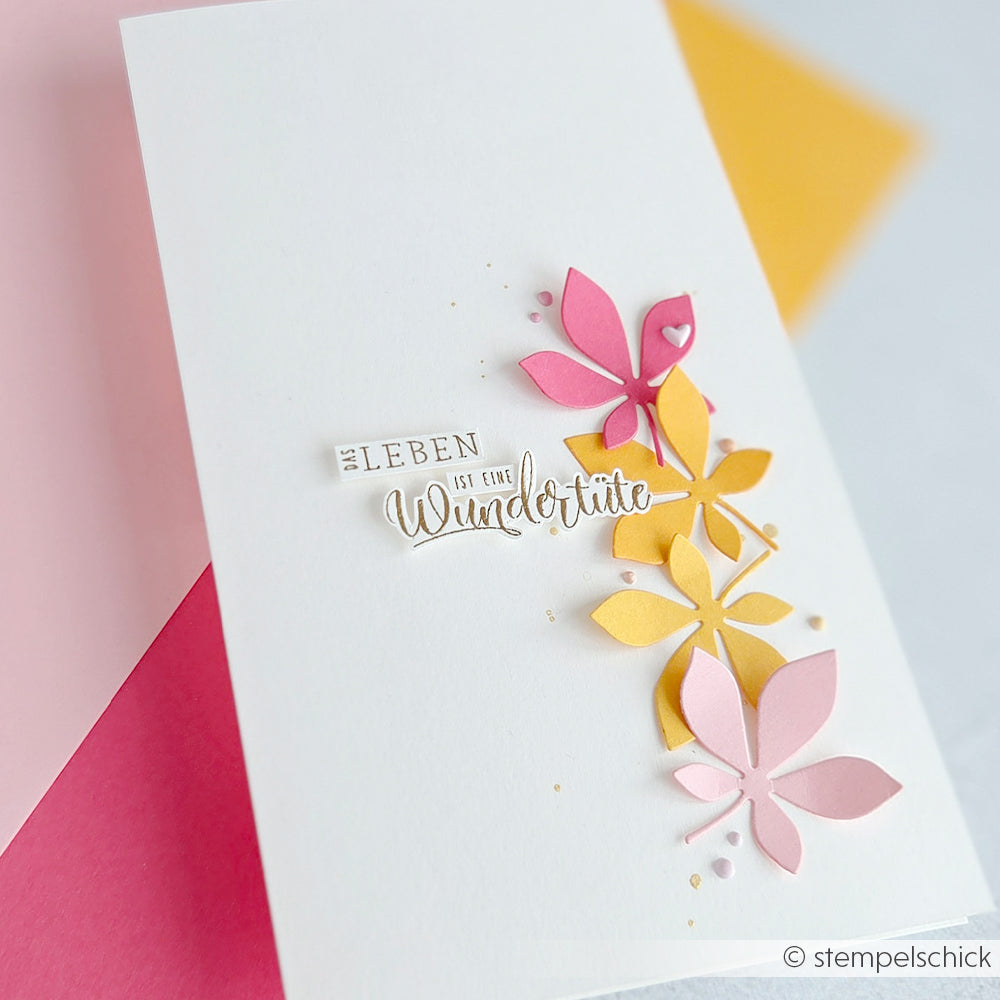 Weiße Karte mit rosa und gelben Papierblumen aus Designpapieren, akzentuiert mit deutschem Text, vor einem rosa-gelben Hintergrund. Mit zarten Details, die mit der Motivstanze „Blätterwirbel“ gestaltet wurden und der Karte einen eleganten Touch verleihen.