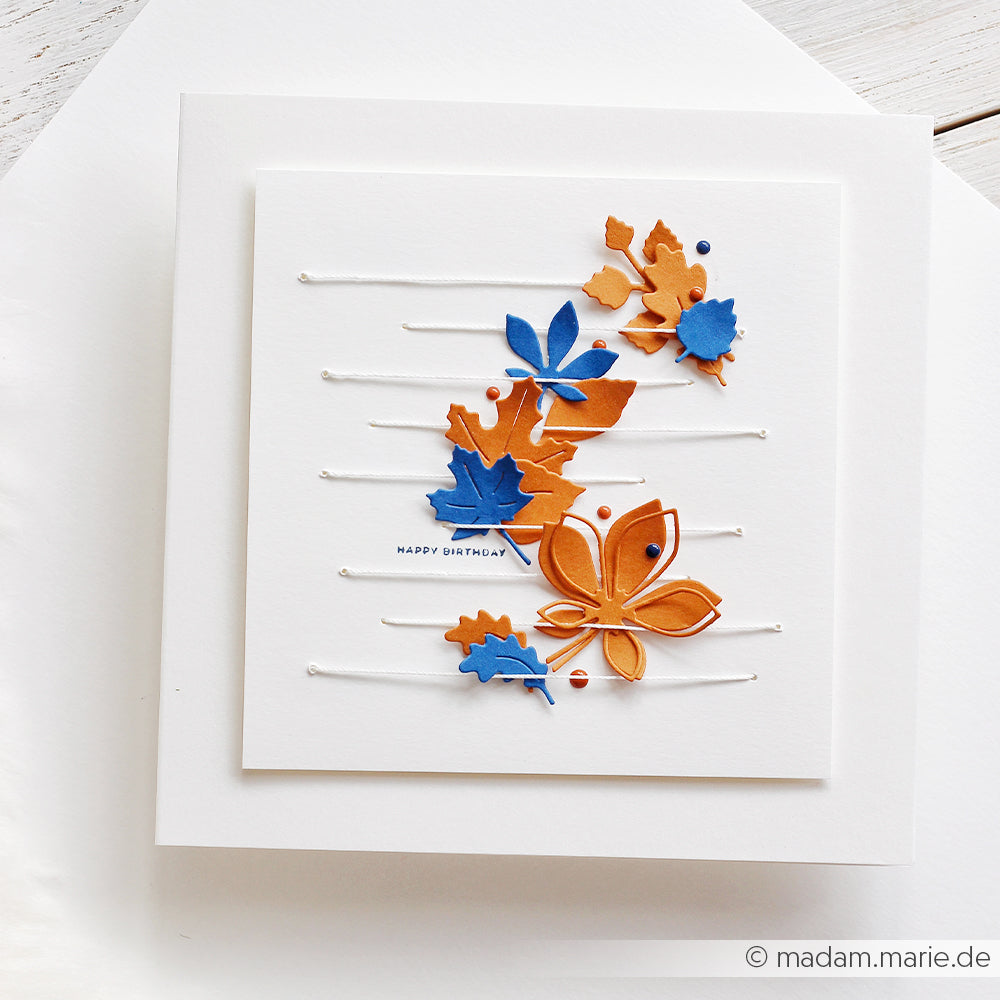 Geburtstagskarte mit blauen und orangefarbenen Papierblättern, gefertigt mit der Motivstanze „Blätterwirbel“ und hochwertigen Designpapieren, zart auf einer weißen Schnur angeordnet, mit dem Schriftzug „Happy Birthday“ auf einem schlichten weißen Hintergrund.