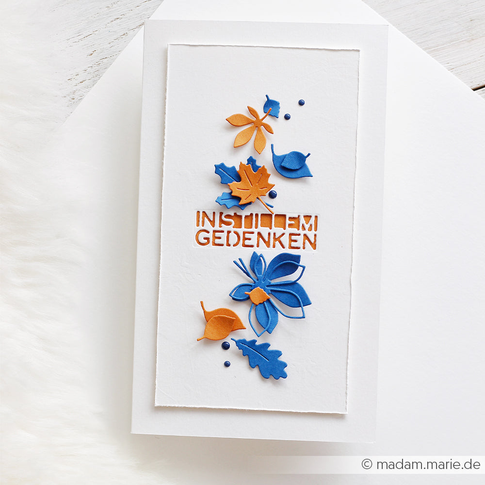 Weiße Karte mit orangefarbenen und blauen Papierblumen und Blättern, gefertigt aus hochwertigen Designpapieren und mit der filigranen Motivstanze „Blätterwirbel“. Mit dem deutschen Text „In stillem Gedenken“.