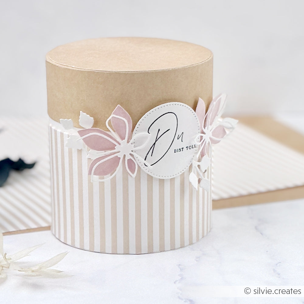 Runde, gestreifte Geschenkbox mit rosa-weißen Papierblumen, die aus edlen Designpapieren mit der Motivstanze „Blätterwirbel“ gefertigt wurden, sowie einem Anhänger mit der Aufschrift „Du bist toll.“ auf weißem Hintergrund.