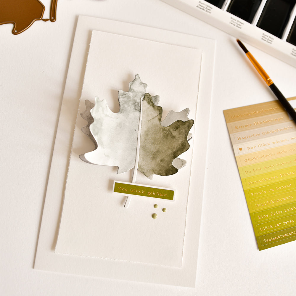 Ein Aquarellbild eines Ahornblatts auf weißem Papier, daneben ein Pinsel und Farbmuster auf einer weißen Fläche – die perfekte Inspiration für Ihr nächstes Projekt mit der Motivstanze „Ahornblatt“ oder passenden Designpapieren.
