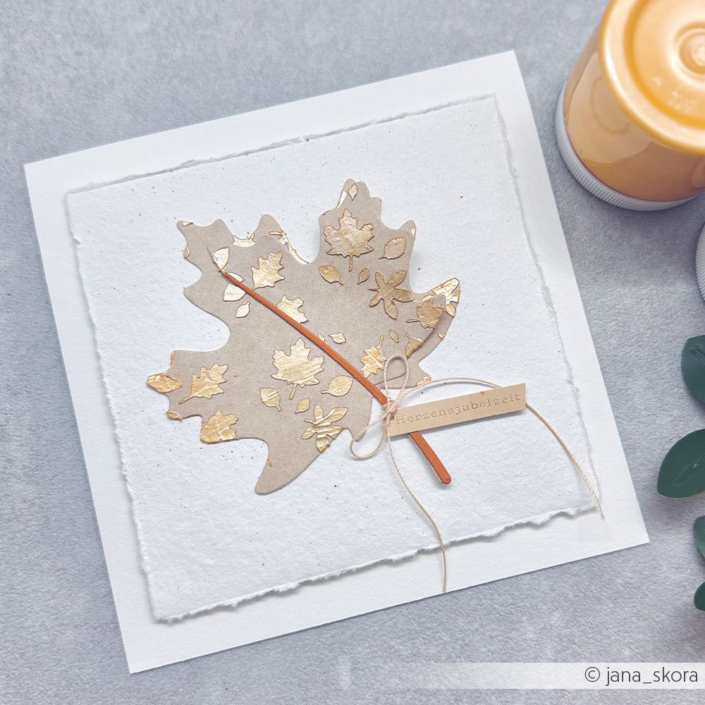 Handgefertigte Karte mit einem Blatt mit goldenen Akzenten, einem Band und einem Anhänger – gestaltet mit der Motivstanze „Ahornblatt“ – präsentiert neben einer gelben Kerze und grünen Blättern. Ideal, um Ihre Lieblings-Designpapiere in Szene zu setzen oder mit kreativen Stanzsystemen zu experimentieren.