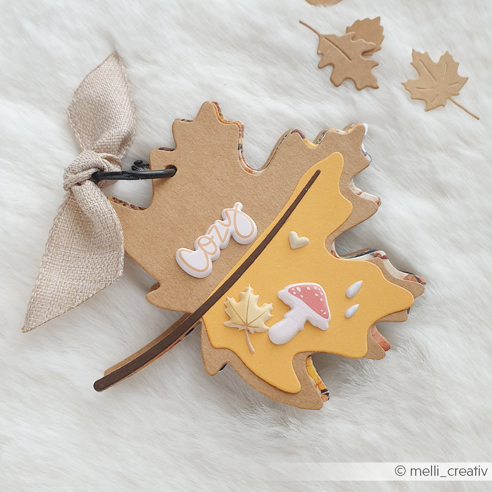 Blattförmige Karte, gefertigt mit der Motivstanze „Ahorn Blatt“ und verziert mit einem gemütlichen Text sowie Akzenten aus Designpapieren mit Herbstmotiven, abgerundet mit einem Band und präsentiert auf einer weißen, flauschigen Unterlage.