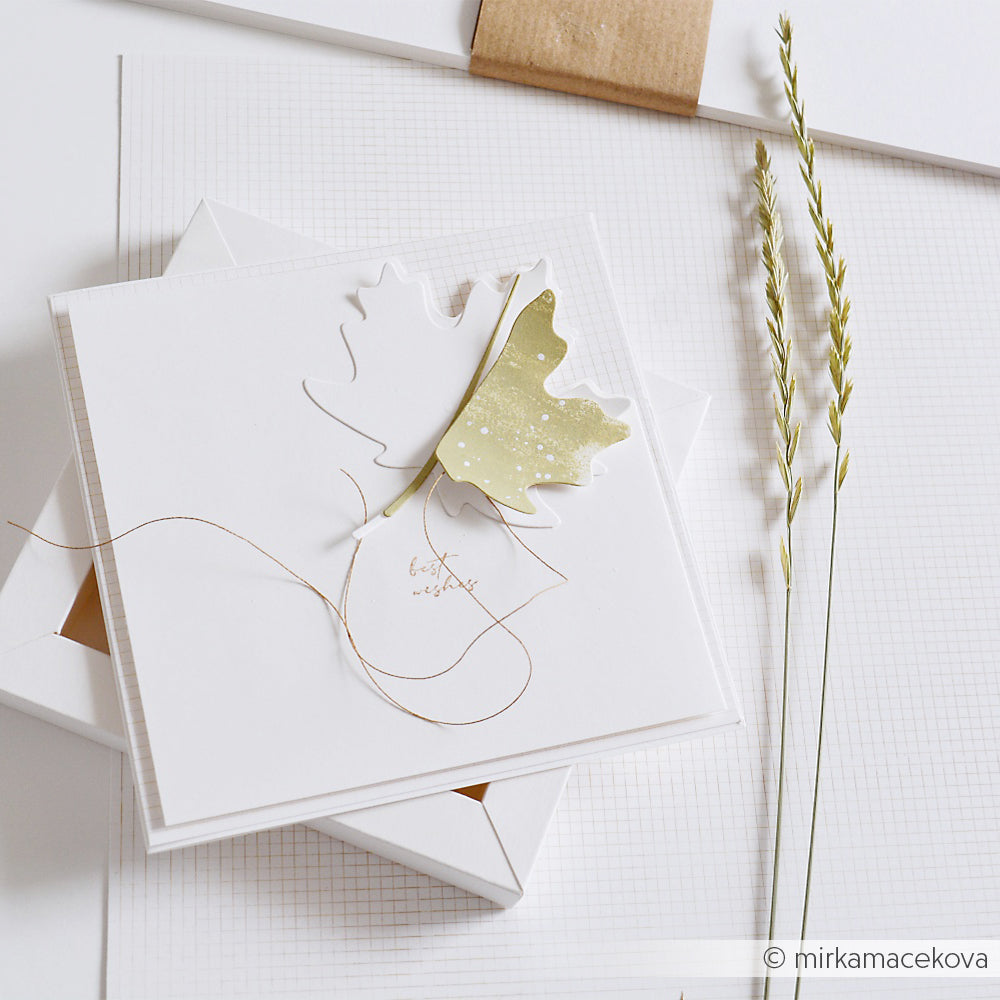 Quadratische weiße Karte mit einer ausgeschnittenen Blattform, gestaltet mit der Motivstanze „Ahorn Blatt Groß“, goldener Schnur und getrocknetem Gras auf einem strukturierten weißen Hintergrund – ideal für kreative Projekte mit Designpapieren und Stanzsystemen.