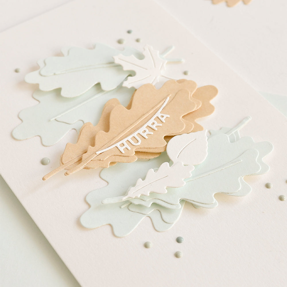 Mehrschichtige Papierblätter in sanften Pastellfarben, gefertigt aus hochwertigen Designpapieren, darunter ein hellbraunes Blatt mit dem Wort „HURRA“ in weißen Buchstaben. Die zarten Formen werden mit der Motivstanze „Eichenblätter“ ausgestanzt, die für klare und elegante Details sorgt.