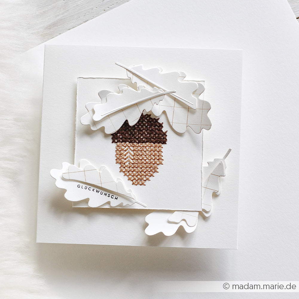 Eine handgefertigte Karte mit einer im Kreuzstich gestickten Eichel und mehrschichtigen Eichenblättern aus Papier, hergestellt aus eleganten Designpapieren und verziert mit zarten Eichenblattformen, die mit Motivstanze-Stanzsystemen ausgestanzt wurden – das Ganze präsentiert auf einem strahlend weißen Hintergrund.