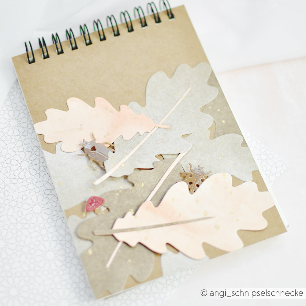 Ein Notizbuch mit pastellfarbenen Eichenblatt-Ausschnitten, hergestellt mit der Motivstanze „Eichenblatt“ und hochwertigen Designpapieren, verziert mit kleinen Tierfiguren aus Holz auf dem Einband.