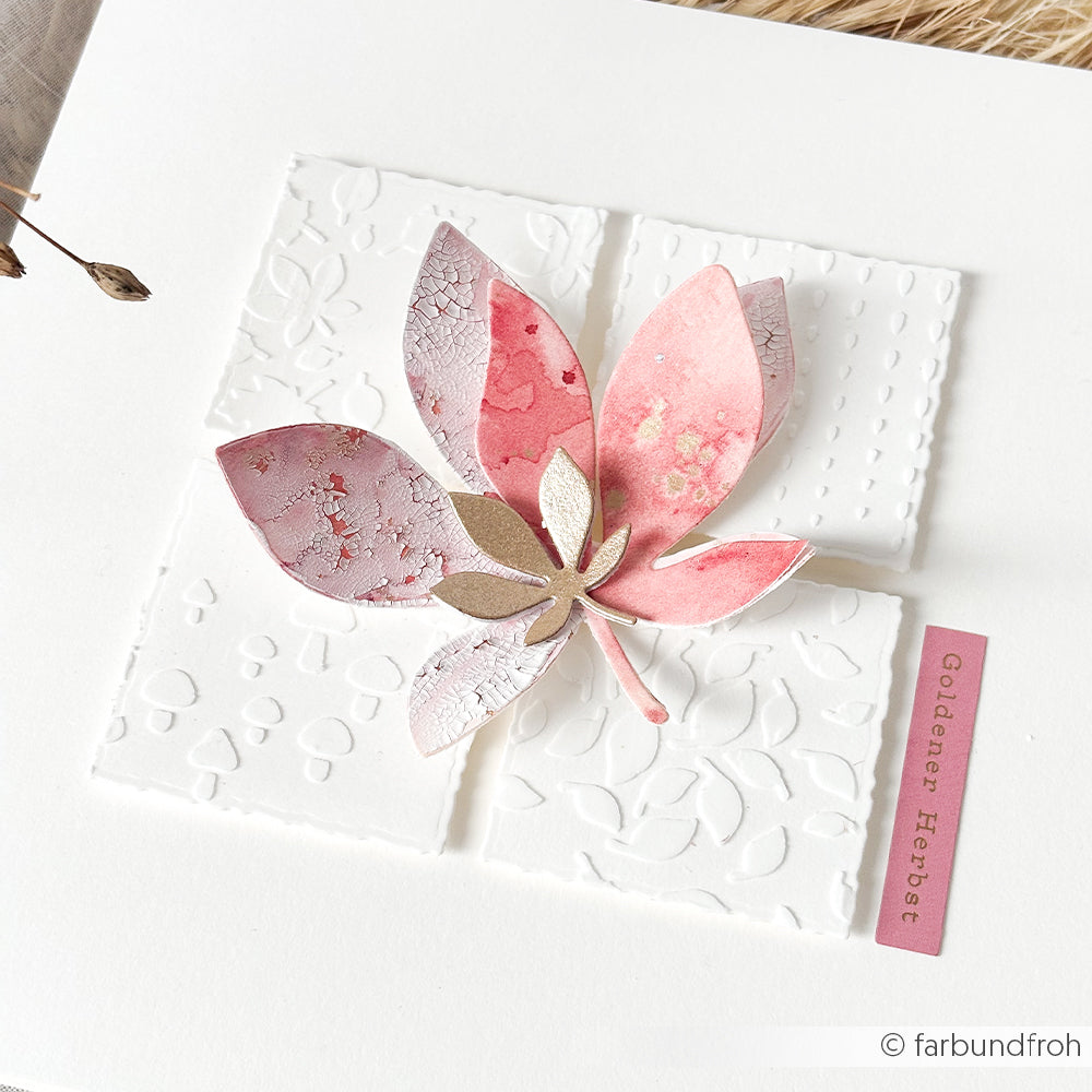 Ein rosa-goldenes Papierblatt, gestaltet mit Designpapieren und der Motivstanze „Kastanienblatt“, ruht auf geprägten weißen Quadraten, auf denen auf einem rosa Etikett „Goldener Herbst“ steht.