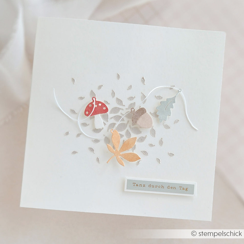 Weiße Karte mit Herbstblättern, einem Pilz, einer Eichel und dem Schriftzug „Tanz durch den Tag“ auf der Vorderseite. Perfekt gestaltet mit der Motivstanze „Herbst-Anhänger“ und den Stanzen von DieCut Systeme für wunderschöne, detailreiche saisonale Akzente.