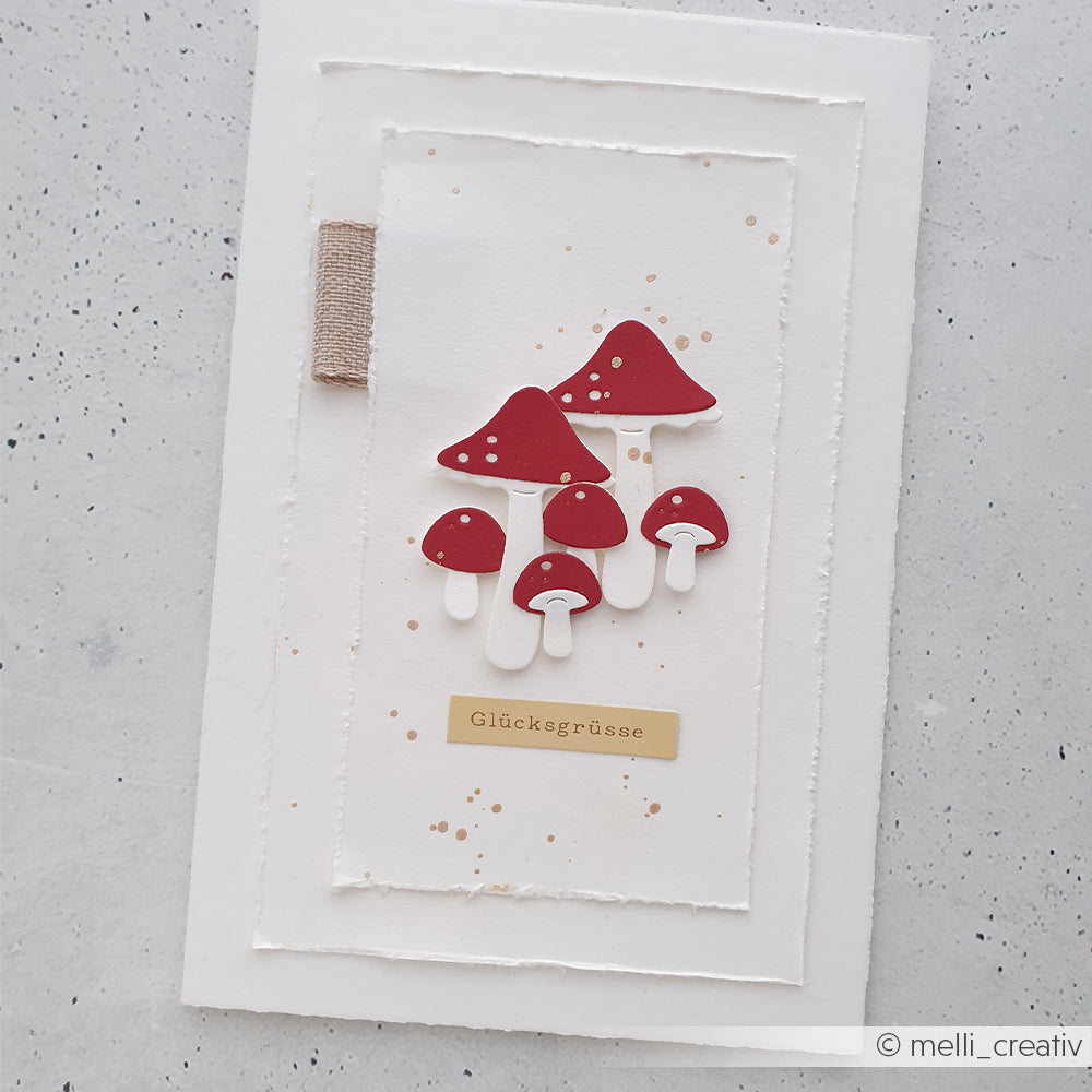 Handgefertigte Karte mit roten Pilzen, gestaltet mit der Motivstanze „Kleines Pilz-Trio“ auf hochwertigem Designpapier, mit dem Schriftzug „Glücksgrüße“ auf weißem Strukturpapier und einem beigen Band auf der linken Seite.
